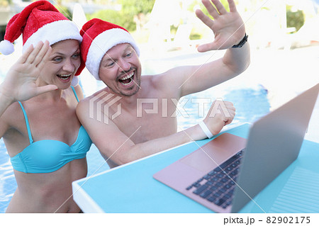 Young man and woman in red santa hats waving at laptop screen on side of pool 82902175