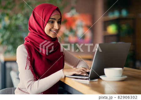 Cheerful middle-eastern woman in hijab using laptop at cafe 82906302