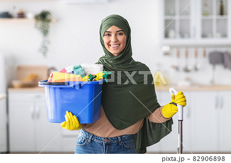 Cheerful muslim housewife holding bucket with cleaning supplies and mop Cheerful muslim housewife holding bucket with cleaning supplies and mop 82906898