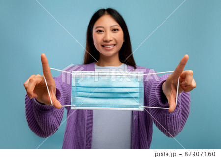 Stop covid concept. Happy asian lady holding medical mask and smiling at camera on blue studio background Stop covid concept. Happy asian lady holding medical mask and smiling at camera on blue studio background 82907160