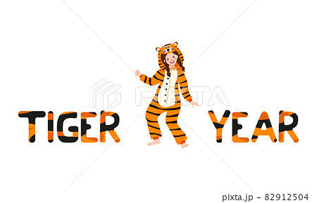 Girl in tiger carnival costume and inscription New Year from striped orange with black letters. Children pajama party or holiday. Kid in jumpsuits or kigurumi, festive clothing for Christmas 82912504