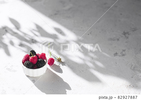 White bowl full with raspberries, blackberry on gray concrete table with leaves, tree branches shadow. Healthy eating concept. Eco, bio farming. Fresh tasty berries on grey background. Quality photo White bowl full with raspberries, blackberry on gray concrete table with leaves, tree branches shadow. Healthy eating concept. Eco, bio farming. Fresh tasty berries on grey background. Quality photo 82927887