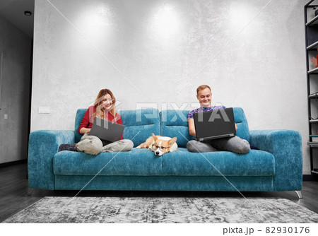 Two smiling people sitting on different ends of blue sofa. Woman invisibly looking into her husband's laptop. Pet dog in glasses is bored, waiting for person to pay attention or play with him Two smiling people sitting on different ends of blue sofa. Woman invisibly looking into her husband's laptop. Pet dog in glasses is bored, waiting for person to pay attention or play with him 82930176