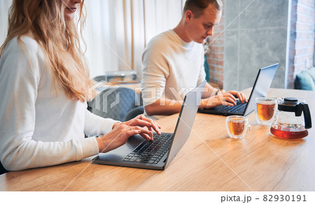 Close up of young woman and man typing on laptop keyboard while spending time together at home. Married couple sitting at wooden table with cups of tea and using modern notebooks. Close up of young woman and man typing on laptop keyboard while spending time together at home. Married couple sitting at wooden table with cups of tea and using modern notebooks. 82930191