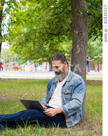 Photo of a bearded man while working online with a laptop. He sits in the park on the grass and works remotely. 82930217