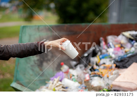 Woman throwing a cardboard glass into a recycling bin. Taking care of the cleanliness of the city and the environment. A large trash bin in a public park Woman throwing a cardboard glass into a recycling bin. Taking care of the cleanliness of the city and the environment. A large trash bin in a public park 82933175
