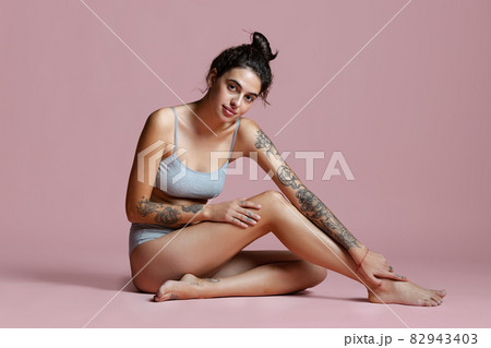 Full-length portrait of beautiful young woman sitting in gray cotton underwear isolated over pink background. 82943403