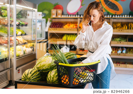 Medium shot of attractive young woman talking on mobile phone and checking shopping list bt shopping basket. 82946266