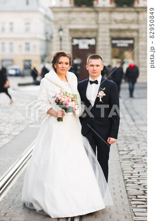 Wedding photo shooting. Bride and bridegroom walking in the city. Married couple embracing and looking at each other. Holding bouquet 82950849