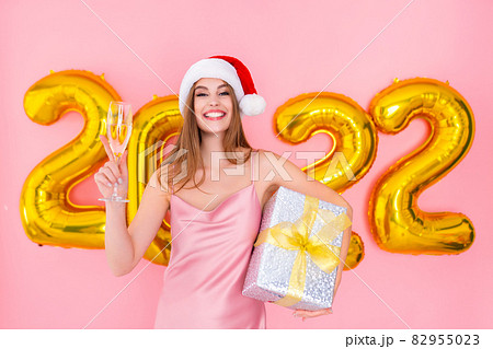 Excited Santa girl raises up glass of champagne while holds gift box. 2022 air balloons New Year Excited Santa girl raises up glass of champagne while holds gift box. 2022 air balloons New Year 82955023
