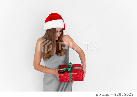 Excited positive girl in Santa's hat shaking gift box while stands on white background. Celebration Excited positive girl in Santa's hat shaking gift box while stands on white background. Celebration 82955025