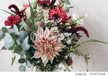 Closeup of colorful flowers. Moody autumn wedding or birthday bouquet. Pink and burgundy dahlia, cosmos and aster flowers. Amaranthus and eucalyptus foliage. Selective focus. White wall. 82973208