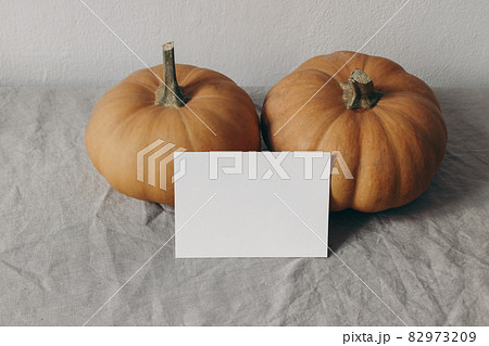 Autumn mockup. Closeup of blank greeting card, invitation with orange pumpkins on beige linen table cloth. Blurred white wall background. Fall stationery. Halloween, Thanksgiving concept. 82973209