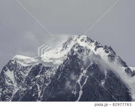 Atmospheric alpine landscape with snow-covered mountain top under snow gray sky. Awesome scenery with beautiful pointy peak with snow and high snowy mountain wall with low clouds. 82977763
