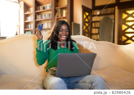 Excited young black lady with credit card and laptop shouting OMG, winning in online lottery, shopping on sale from home 82980426