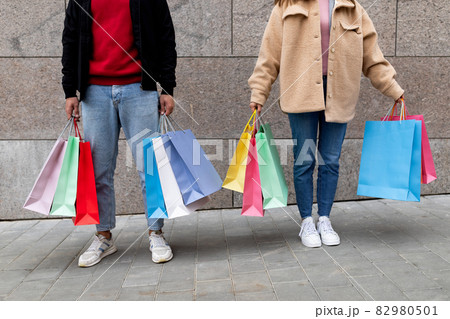 Unrecognizable young couple holding shopper bags, posing in front of stone wall outdoors, going shopping together Unrecognizable young couple holding shopper bags, posing in front of stone wall outdoors, going shopping together 82980501