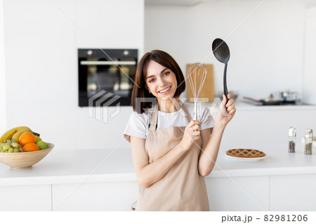 Portrait of happy lady wearing apron and posing in kitchen holding kitchenware utensils in hands and smiling at camera 82981206