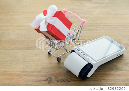 Closeup of credit card terminal with touchscreen display and red gift box with white bow in miniature shopping cart on a wooden table Closeup of credit card terminal with touchscreen display and red gift box with white bow in miniature shopping cart on a wooden table 82982202