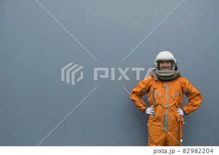 Astronaut wearing orange space suit and space helmet smiling while posing against gray wall background 82982204