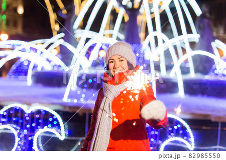 Smiling young woman wearing winter knitted clothes holding sparkler outdoors over snow background. Christmas holidays. 82985550