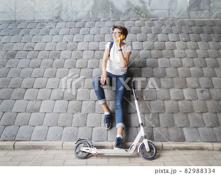 Smiling woman talks by smartphone after riding kick scooter. Female in jeans and sneakers on grey tiled wall background. Healthy lifestyle. Eco-friendly urban transport. 82988334