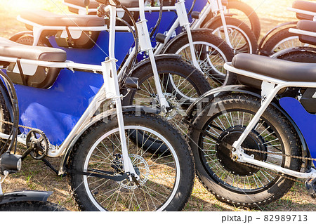 Many retro fat electric bicycle scooters parked on dirt grass field outdoor ready for hire rental sharing cpuntry parl leisure recreation trip. E power transport rent equipment. Rural offroad cycling 82989713