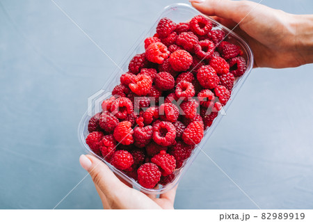 Fresh ripe raspberries in plastic box in hands on blue background, close-up. Fresh ripe raspberries in plastic box in hands on blue background, close-up. 82989919