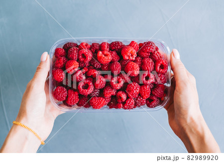 Fresh ripe raspberries in plastic box in hands on blue background, close-up. Fresh ripe raspberries in plastic box in hands on blue background, close-up. 82989920