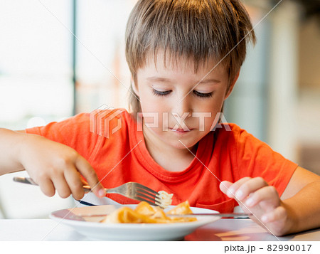 Cute kid in red t-shirt eats pancakes with knife and fork. Curious boy with funny expression on face. Tasty pastry for breakfast. 82990017