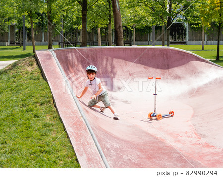 Little boy fell off kick scooter while riding in skate park. Special concrete bowl structures in urban park. Training to skate at summer. 82990294