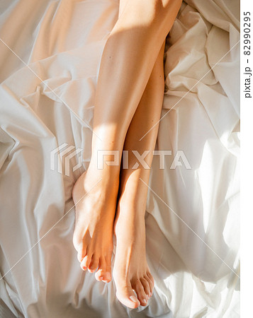 Morning at bed. Woman's feet on unmade bed. Sunbeams on white crumpled bed sheet. Light and shadow. 82990295