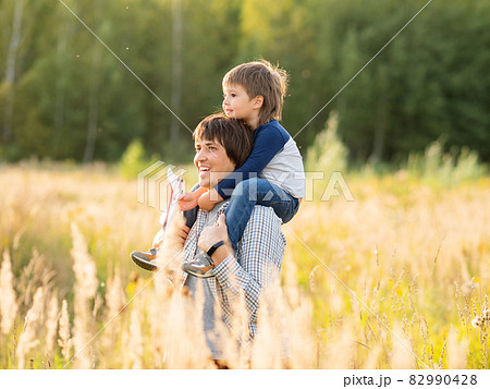 Father carries his son on shoulders. Family time outdoors. Man with child on autumn field at sunset. Dad and son explores nature together. 82990428
