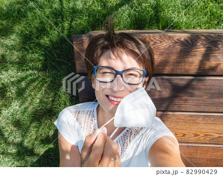 Smiling woman takes off medical protective mask to make selfie on her smartphone. Female in eyeglasses has a rest in urban park. New normal. Coronavirus COVID-19 quarantine and precautional measures. 82990429