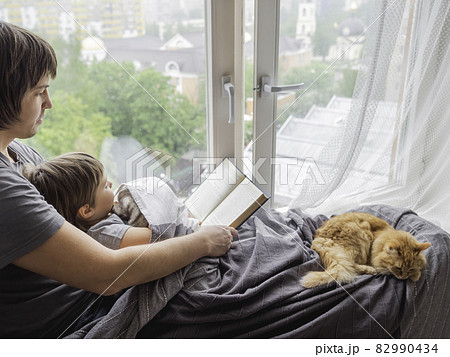 Father reads book to his son. Cozy family time on window sill with sleepy ginger cat. Dad, toddler boy and fluffy pet at home. 82990434