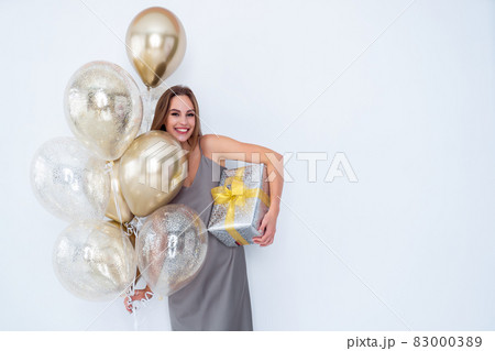 Photo of smiling girl holds large wrapped giftbox and many air balloons. came to party. Celebration  83000389