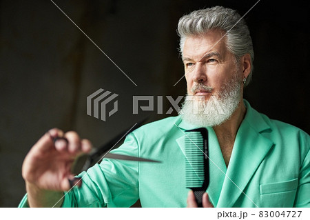 Portrait of stylish gray haired mature man with beard wearing cool outfit looking away, holding sharp barber scissors and hair comb, standing over dark background 83004727