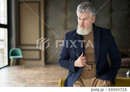 Stylish handsome mature man in casual clothes looking away while posing, leaning on sofa indoors Stylish handsome mature man in casual clothes looking away while posing, leaning on sofa indoors 83004728