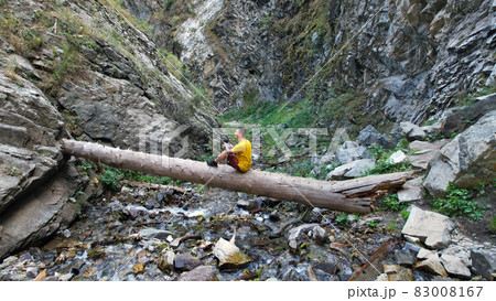 The guy is sitting on a big tree above the river. 83008167
