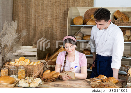 An attractive new saleswoman describes wooden plates with prices for individual types of bread under the supervision of a bakery owner, a young handsome baker. An attractive new saleswoman describes wooden plates with prices for individual types of bread under the supervision of a bakery owner, a young handsome baker. 83014301
