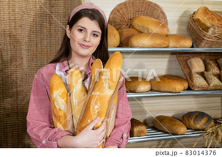 A beautiful saleswoman hugs long baguette rolls in her arms and intends to serve them to the customer. Sale of bread in a traditional bakery store. A beautiful saleswoman hugs long baguette rolls in her arms and intends to serve them to the customer. Sale of bread in a traditional bakery store. 83014376