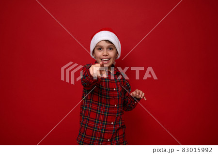 Adorable preadolescent boy in red checkered shirt wearing a Santa hat points directly to camera, smiles with beautiful toothy smile posing against colored background with copy space for Christmas ad 83015992