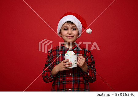 Portrait of amazing preteen cute boy, adorable child in Santa hatand plaid shirt holding a mug ith hot chocolate with marshmallows, smiles looking at camera against red background with copy space 83016246