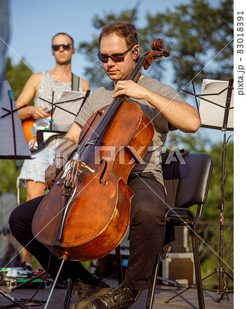 Male violoncellist playing cello in orchestra outdoors Male violoncellist playing cello in orchestra outdoors 83018391