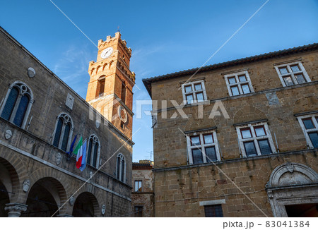 Pienza, Tuscany, Italy. August 2020. The late afternoon sun illuminates the clock tower in the historic center with warm light. Below the flags on the facade of the town hall. 83041384