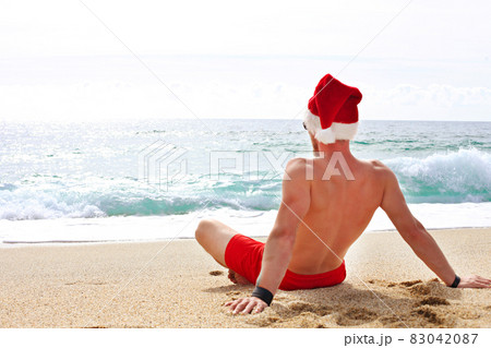Muscle male in santa claus hat and red swimsuit, lying on sand, relaxing on sea shore. Christmas in tropical destinations. 83042087