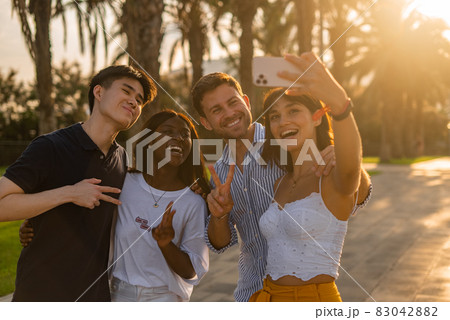 Company of joyful young multiracial people making peace gesture and taking selfie on smartphone while enjoying summer holidays together in tropical city Company of joyful young multiracial people making peace gesture and taking selfie on smartphone while enjoying summer holidays together in tropical city 83042882