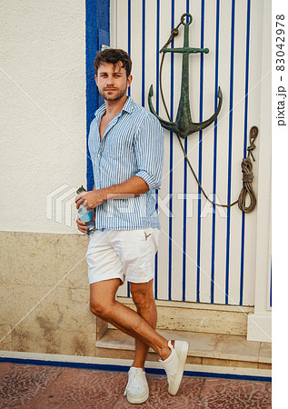 Full body adult man with bottle of water leaning on wall near door decorated with anchor in summer 83042978
