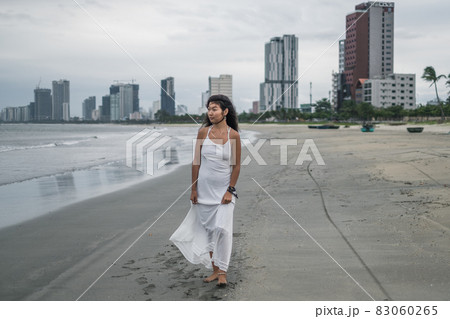 Pensive young asian woman in white dress standing on the beach and looking at the ocean. Romantic and peaceful photo. High quality photo 83060265