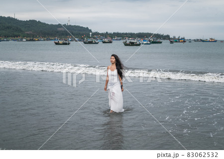 Charming young mongol woman in white dress walking on the beach. Black long curly hair. Looking aside. Romantic photo. Ocean view. High quality photo 83062532