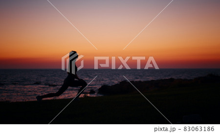 Silhouette of active young woman in sport clothes stretching arms and legs during amazing sunset over beach. Concept of people, wellness and body care. Silhouette of active young woman in sport clothes stretching arms and legs during amazing sunset over beach. Concept of people, wellness and body care. 83063186